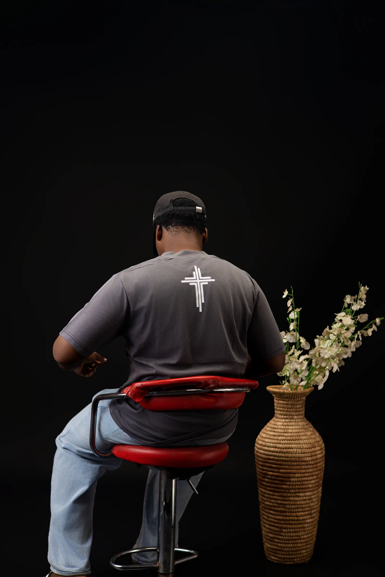 Male model seated with his back to camera, showing the white cross print on the dark grey shirt