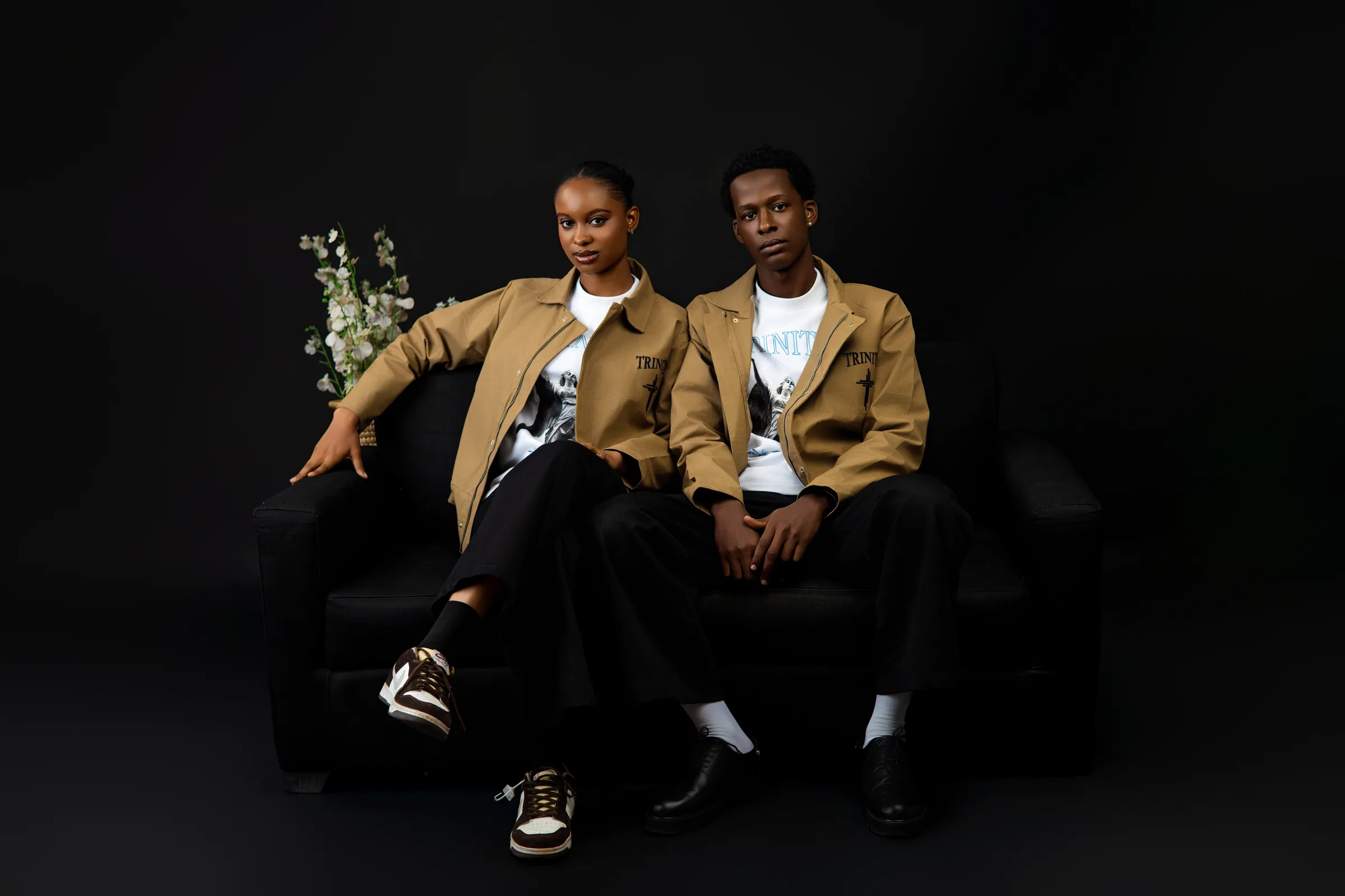 Both models seated together, wearing tan jackets over white graphic shirts and black pants
